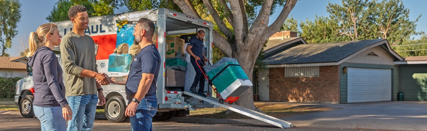 A couple talks to a Moving Helper while additional Moving Helpers load a washing machine into a U-Haul truck in the background.